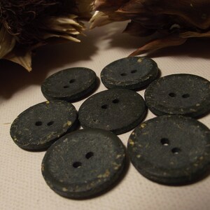 May include: Seven dark grey buttons with two holes each. The buttons are round and oval, with a textured surface and flecks of yellow. They are arranged on a light beige surface, with dried plant matter in the background.