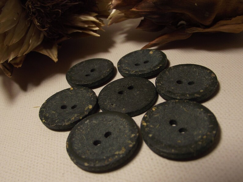 May include: Seven dark grey buttons with two holes each. The buttons are round and oval, with a textured surface and flecks of yellow. They are arranged on a light beige surface, with dried plant matter in the background.