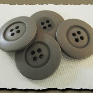 Puede incluir: Cuatro botones grandes y redondos de color topo, cada uno con cuatro agujeros. Los botones tienen un borde circular en relieve alrededor de los agujeros centrales. Los botones están dispuestos ligeramente superpuestos sobre un fondo blanco.