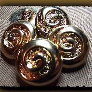 Könnte beinhalten: Eine Sammlung goldfarbener Knöpfe mit einem Spiraldesign. Die Knöpfe haben eine glänzende Oberfläche und sind auf einer strukturierten, neutralfarbenen Oberfläche angeordnet. Die Knöpfe sind wahrscheinlich zum Nähen oder Basteln.