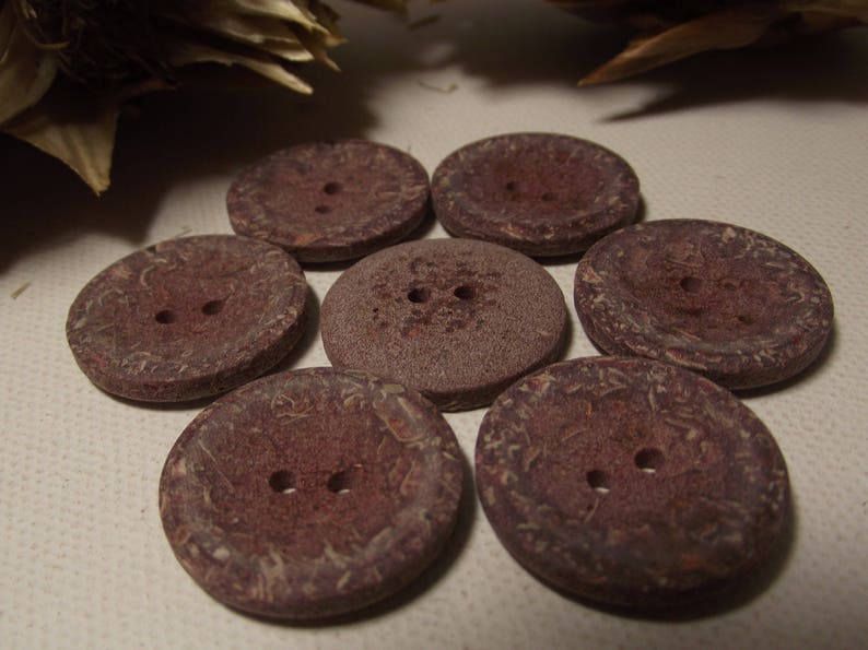 May include: Seven circular, reddish-brown buttons, each with two holes. The buttons display a textured surface and varying shades of brown, possibly made from a natural material. Suitable for sewing and craft activities.