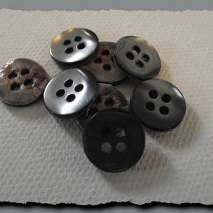 May include: A selection of buttons in grey and black hues. The buttons feature four holes and a slightly curved design. Some have a glossy, reflective appearance, while others have a more matte finish. The buttons are arranged on a textured, off-white surface.