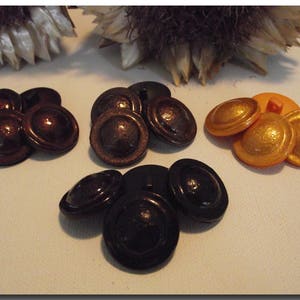 May include: A collection of vintage buttons in various colors. The buttons are in shades of brown, black, and gold. The buttons have a domed shape with a textured surface. The buttons are arranged on a white surface.