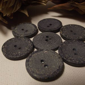 May include: A close-up shot of seven dark grey, round buttons, each with two holes. The buttons are arranged in a circular pattern. They have a textured surface and a slightly weathered look, suggesting a vintage or rustic style.