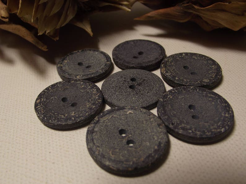 May include: A close-up shot of seven dark grey, round buttons, each with two holes. The buttons are arranged in a circular pattern. They have a textured surface and a slightly weathered look, suggesting a vintage or rustic style.