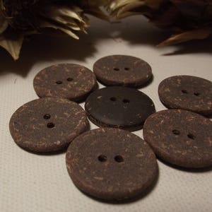 May include: Seven round, brown buttons with two holes each. The buttons are made of a textured material, with one button in a darker shade of brown. The buttons are arranged in a circular pattern.