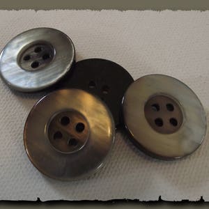 Puede incluir: Un conjunto de cinco botones redondos en varios tonos de marrón y gris. Cada botón tiene cuatro agujeros y un ligero brillo iridiscente. Los botones están dispuestos sobre una superficie texturizada de color blanquecino.