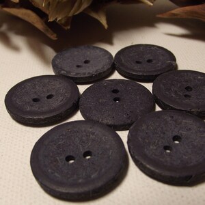 May include: Seven dark grey, round buttons, each with two holes, arranged in a circular pattern. The buttons have a textured surface. Suitable for sewing or craft projects.