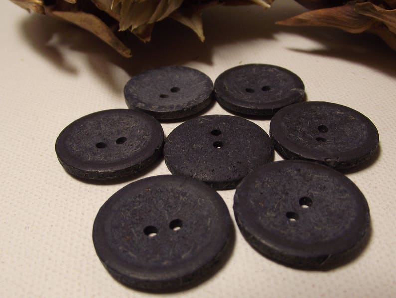 May include: Seven dark grey, round buttons, each with two holes, arranged in a circular pattern. The buttons have a textured surface. Suitable for sewing or craft projects.