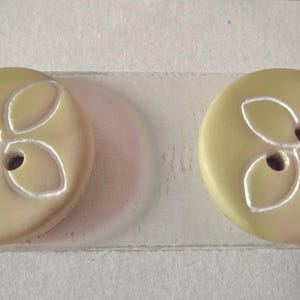 May include: Two round, pale yellow buttons with a floral design. Each button has two holes and a white, leaf-like pattern. The buttons are made of a smooth material and are suitable for sewing projects.