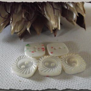 Peut inclure: Cinq boutons blanc cassé sont présentés. Deux ont un motif en spirale, et trois sont carrés avec deux ou trois trous. Certains boutons ont de petites mouchetures rouges et vertes. Les boutons sont sur une surface blanche texturée.