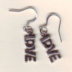 Puede incluir: Un par de pendientes plateados. Cada pendiente deletrea la palabra "LOVE" en mayúsculas. Los pendientes tienen un diseño de gancho para orejas perforadas. El fondo es de color beige claro.