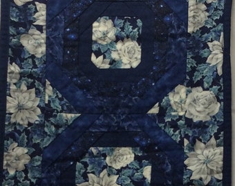 May include: A blue and white quilted table runner with a geometric design featuring white poinsettia flowers and a dark blue background. The runner has a diamond shape with a pointed end.