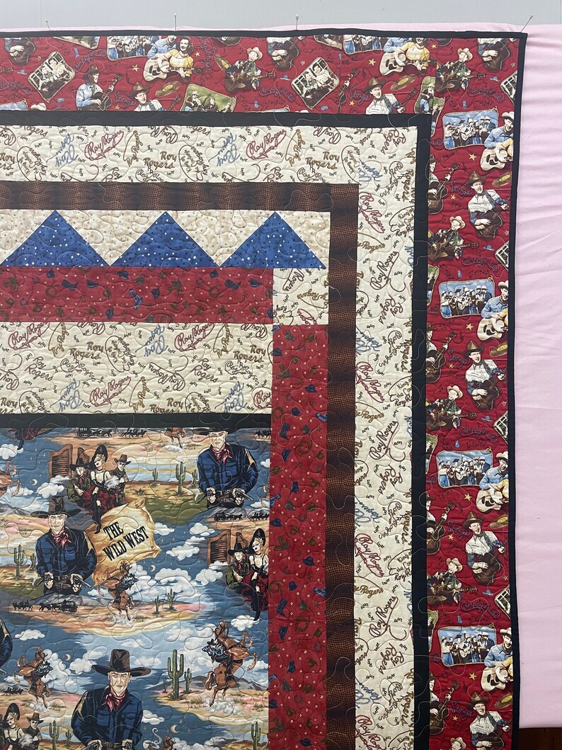 May include: A patchwork quilt featuring Roy Rogers themed fabric with western motifs. The design includes panels of cowboys, desert landscapes with cacti, and text reading The Wild West. The quilt is bordered by red and cream patterned fabric.