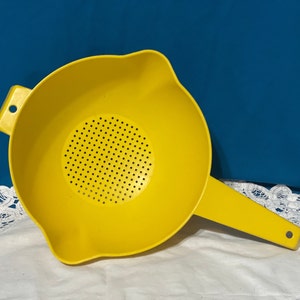 May include: Bright yellow plastic colander with a handle and a perforated straining surface. The colander has a pouring spout and a small hole on the handle for hanging. The kitchen utensil is set against a blue background.