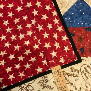 May include: A quilted fabric piece featuring a red background with white stars and a black border, layered over a cream-colored fabric with Roy Rogers text and rope patterns. A wooden ruler shows measurements starting from 11 to 15 inches.
