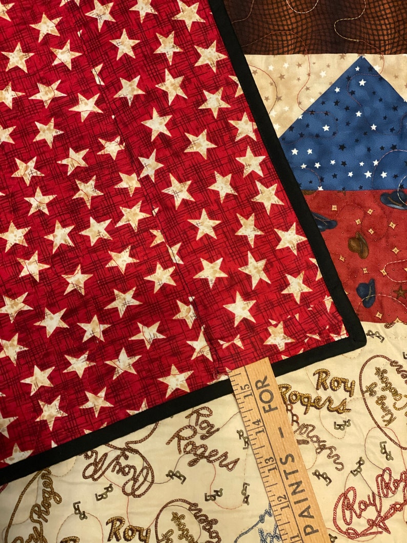 May include: A quilted fabric piece featuring a red background with white stars and a black border, layered over a cream-colored fabric with Roy Rogers text and rope patterns. A wooden ruler shows measurements starting from 11 to 15 inches.