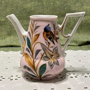 May include: A pink ceramic pitcher with a bird and floral design. The pitcher has a curved spout and a handle with a geometric design. The bird is perched on a branch with leaves and flowers. Gold accents are on the handle and spout.