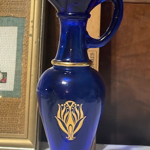 May include: A cobalt blue glass bottle with a gold trim and a gold floral design. The bottle has a spout and a handle.