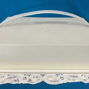 May include: White plastic cake carrier with a clear lid and a handle. The carrier is sitting on a white lace doily.