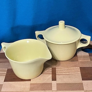 May include: A vintage pale yellow creamer and sugar bowl set with a lid. The creamer has a curved spout and handle. The sugar bowl has a lid with a small handle and two side handles. The set is on a wooden cutting board.
