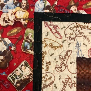 May include: A quilted fabric featuring a red section with printed illustrations of Roy Rogers playing guitar, singing, and wearing cowboy hats, alongside a cream-colored section with repeating cursive text reading Roy Rogers and lasso motifs.