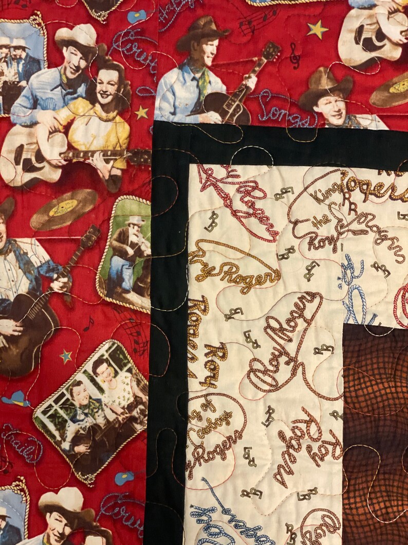 May include: A quilted fabric featuring a red section with printed illustrations of Roy Rogers playing guitar, singing, and wearing cowboy hats, alongside a cream-colored section with repeating cursive text reading Roy Rogers and lasso motifs.
