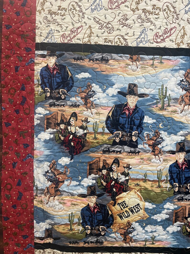 May include: A quilted fabric featuring a Wild West theme with repeating illustrations of Roy Rogers, cowboys, and cowgirls in desert landscapes. The design includes cacti, horses, and a scroll with the text THE WILD WEST, bordered by a red patterned strip.