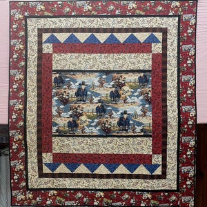 May include: A rectangular quilt featuring a central panel with cowboys, cacti, and the text The Wild West. The quilt is bordered by layers of cream fabric with rope patterns, red fabric with small motifs, and blue triangles on a cream background.