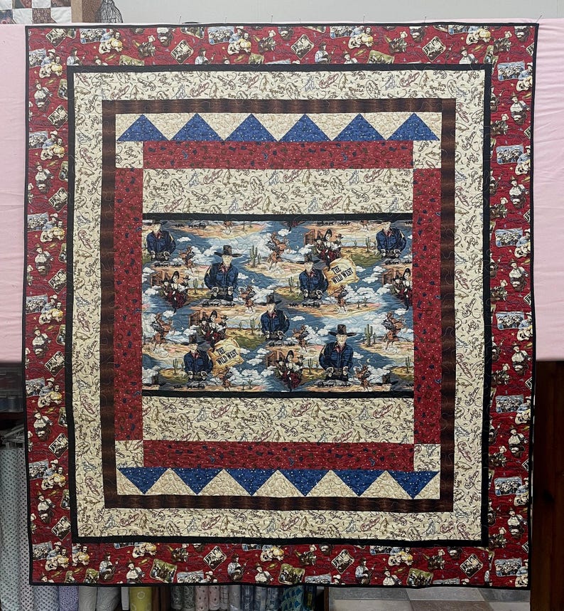May include: A rectangular quilt featuring a central panel with cowboys, cacti, and the text The Wild West. The quilt is bordered by layers of cream fabric with rope patterns, red fabric with small motifs, and blue triangles on a cream background.