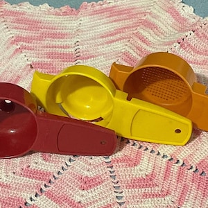 May include: Three vintage kitchen tools in red, yellow, and orange. The red tool is a scoop, the yellow one has a bowl shape, and the orange one has a strainer. They are arranged on a pink and white crocheted blanket.