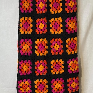 May include: A black crochet skirt featuring a repeating pattern of colorful granny squares. The squares are in shades of orange, pink, and yellow. The skirt has a simple, elastic waistband.