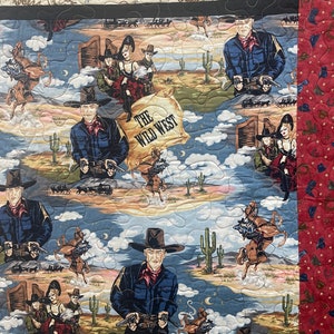 May include: A quilted fabric featuring a Western theme with illustrations of cowboys, horses, and cacti. The central design includes the text "THE WILD WEST" on a parchment-style background. The border is a deep red fabric with small icons.