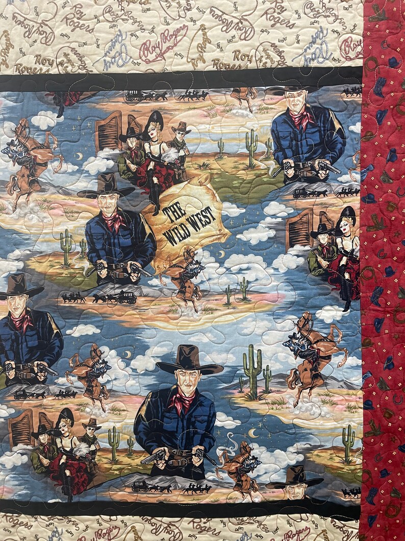May include: A quilted fabric featuring a Western theme with illustrations of cowboys, horses, and cacti. The central design includes the text "THE WILD WEST" on a parchment-style background. The border is a deep red fabric with small icons.