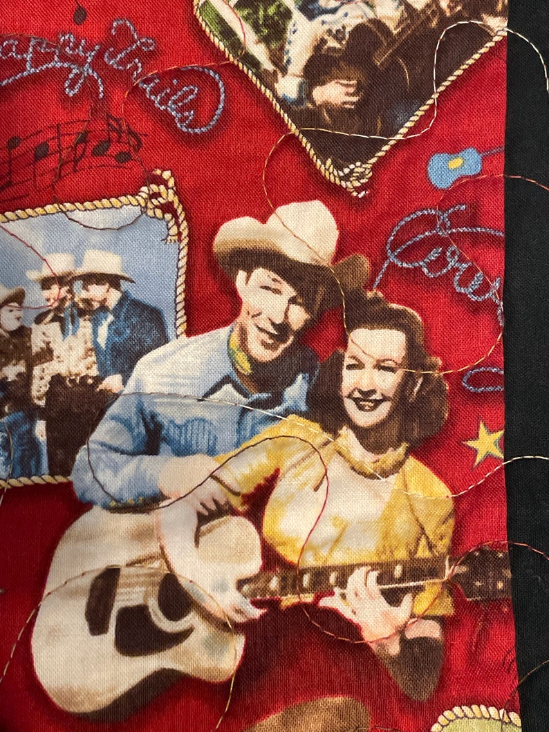 May include: A red fabric featuring a vintage-style print of a cowboy and cowgirl playing a guitar, with additional imagery of musical notes, a rope-bordered photo of people in cowboy hats, and the embroidered text Happy Trails in blue thread.