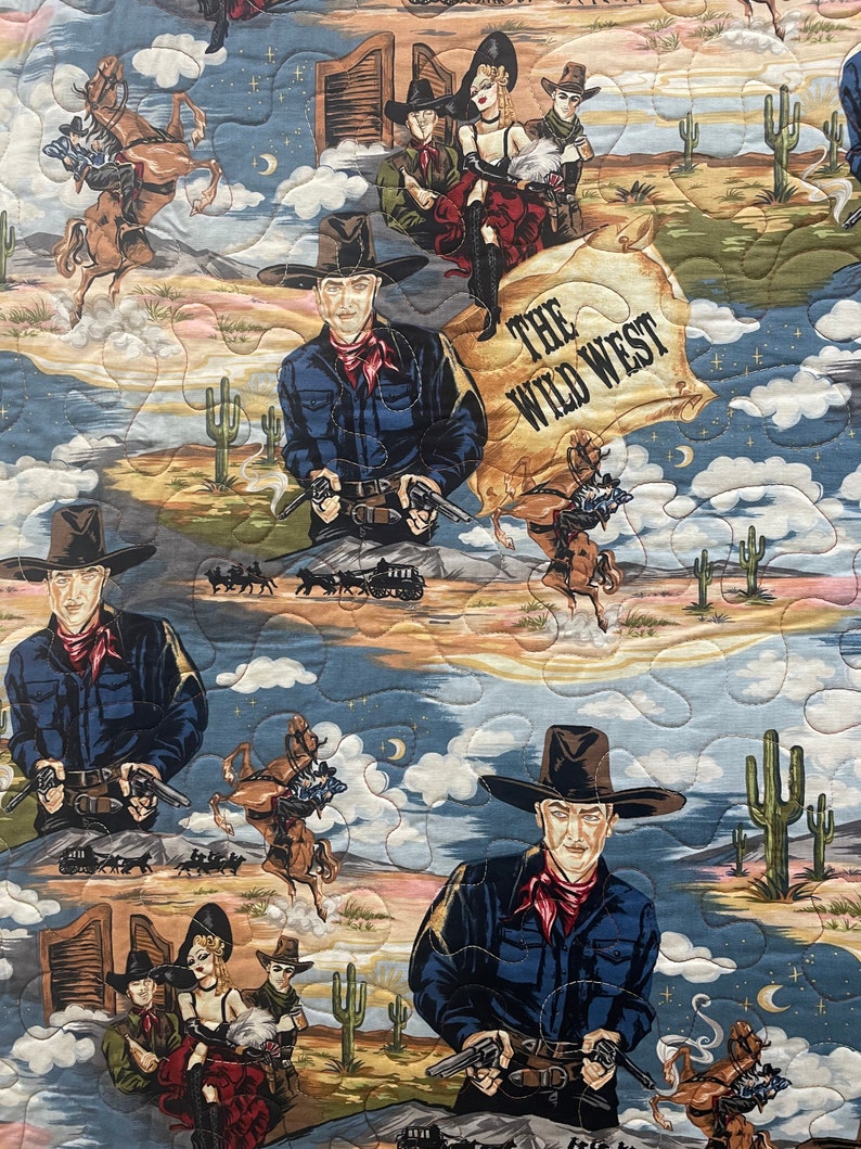 May include: A quilted fabric featuring a Wild West theme with illustrations of cowboys in dark blue shirts and red neckerchiefs, saloon performers, horses, cacti, and desert landscapes. The text THE WILD WEST is printed on a parchment-style scroll.