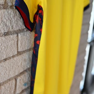 May include: A bright yellow athletic shirt featuring a dark blue side panel with a red abstract splatter pattern. The sleeve cuffs are finished with a dark blue trim, and the fabric appears smooth and lightweight, designed for active movement.