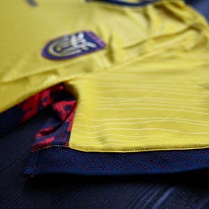 May include: A yellow athletic jersey featuring thin horizontal lines, a dark blue ribbed sleeve cuff with a subtle chevron pattern, and a side panel displaying a red and dark blue floral graphic print. A team crest is visible on the chest area.