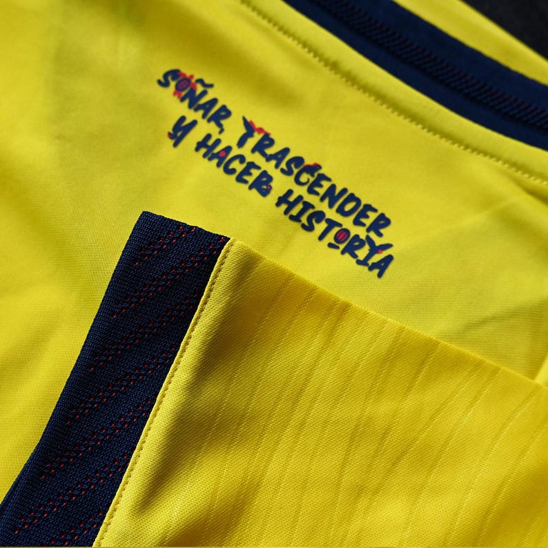 May include: A bright yellow athletic shirt featuring a dark blue ribbed collar and sleeve trim. The fabric displays a subtle diagonal texture. Printed on the chest in dark blue text is the phrase: SO&Ntilde;AR, TRASCENDER Y HACER HISTORIA.