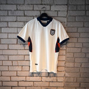 May include: White football shirt with navy blue trim on the collar and sleeves. The shirt has a crest on the chest and a patterned design on the sides. It is hanging on a black hanger against a brick wall.