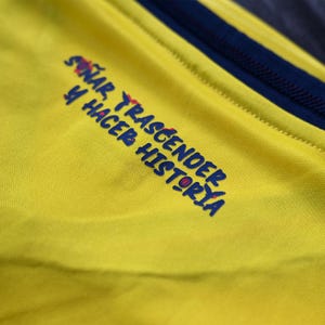 May include: A bright yellow athletic shirt featuring a dark blue collar and the text "SO&Ntilde;AR, TRASCENDER Y HACER HISTORIA" printed in dark blue with small red accents on the chest area. The fabric has a fine, textured, moisture-wicking appearance.