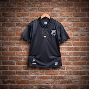 Puede incluir: Polo negro de manga corta, colgado de una percha de madera contra una pared de ladrillo. La camisa presenta un emblema blanco en el pecho y un pequeño detalle tricolor. La camisa tiene cuello y tapeta abotonada.