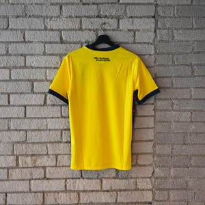 May include: A bright yellow short-sleeve jersey with dark blue trim on the collar and sleeve cuffs. The back features the black text "SO&Ntilde;AR, TRASCENDER Y HACER HISTORIA" printed near the neckline. The shirt is displayed on a black hanger.