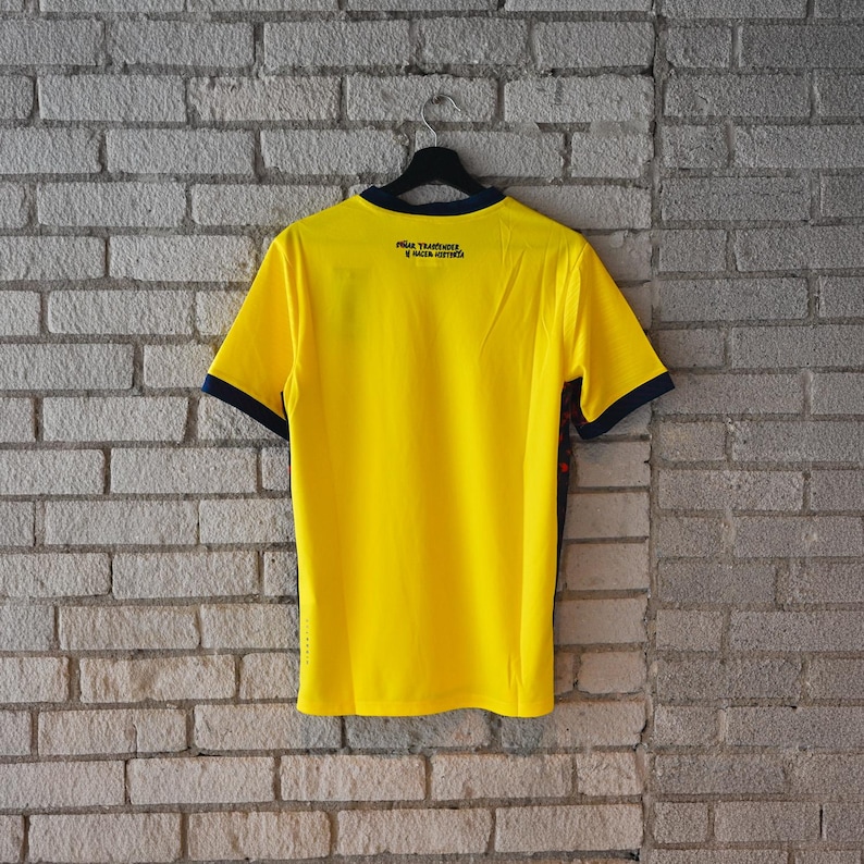 May include: A bright yellow short-sleeve jersey with dark blue trim on the collar and sleeve cuffs. The back features the black text "SO&Ntilde;AR, TRASCENDER Y HACER HISTORIA" printed near the neckline. The shirt is displayed on a black hanger.