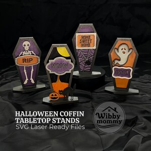 May include: Four wooden coffin-shaped tabletop stands featuring Halloween designs. One shows a skeleton with the text RIP, one a cat and pumpkin with MEOW, one a spiderweb with HOME SWEET HOME, and one a ghost with BOO. The text Wibby mommy is visible.