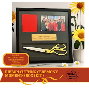 May include: A black shadow box frame containing a red ribbon, a photograph of six people, a gold-colored nameplate with the text RLL Consulting Ribbon Cutting - December 25, 2023, and a pair of large gold-handled scissors with silver blades.