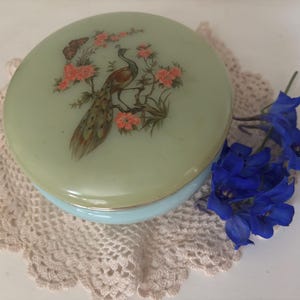 May include: A vintage powder compact with a pale green lid, decorated with a peacock and floral motif. The compact rests on a cream-coloured doily, with a bunch of blue flowers nearby. The base of the compact is light blue.