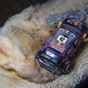 Puede incluir: Un modelo en miniatura de un coche de carreras Red Bull, número 42, con un diseño morado y negro, se encuentra en un diorama de una pista de tierra. El modelo tiene la bandera irlandesa. El diorama incluye arena, rocas y una superficie texturizada.