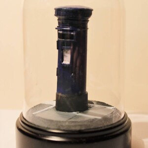 May include: A miniature blue post box under a glass dome on a black base. The post box has a small window and a white painted stripe.
