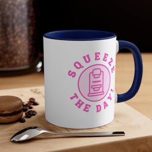 May include: White ceramic coffee mug with a blue rim and handle. The mug has a pink circle with a coffee maker graphic and the text "Squeeze the Day".